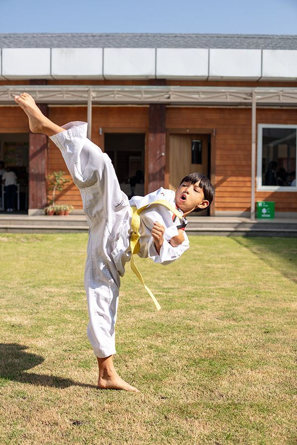 A student at Imperial World School practicing taekwondo outdoors, demonstrating a high kick in a white uniform.